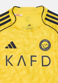 Gele voetbalshirt met een gemusterde textuur, voorzien van marineblauwe strepen op de schouders, de tekst "KAFD" en een ronde logo met "NASSR" en "RIYADH 1965."