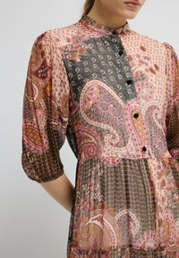 Šaty s mnohobarevným paisley vzorem, vysokým límcem, knoflíkovou přední částí a nařasenými rukávy. Lehká, splývavá tkanina s různými texturami.