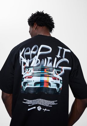 Črna majica s potiskom dirkalnega avtomobila na hrbtni strani in napisom "KEEP IT BURNING" v belem fontu, ki spominja na napis s pršilno barvo.