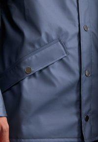 Veste imperméable bleu marine avec une poche à rabat, fermeture à bouton-pression et texture lisse et brillante.