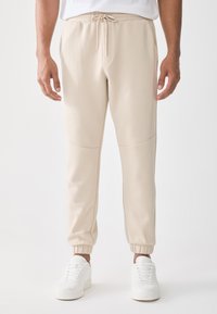 Mann iført beige joggebukser med snor i livet og elastiske mansjetter, kombinert med hvite joggesko, stående mot en ensfarget hvit bakgrunn.