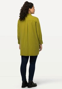 Blusa de manga larga en verde oliva con cuello, detalle de pliegue en la espalda y puños abotonados, combinada con jeans oscuros y botas negras hasta el tobillo.