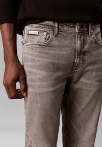 Gros plan d'une personne portant un jean gris Calvin Klein avec un haut noir, la main gauche détendue le long du corps.