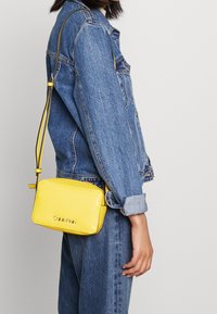 Borsa a tracolla in pelle gialla con forma rettangolare, texture liscia e marchio discreto. Tracolla regolabile attaccata a una chiusura con zip.