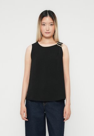Ung kvinne med langt, rett hår iført en ermeløs svart topp med gullspenne og mørkeblå jeans, stående mot hvit bakgrunn.