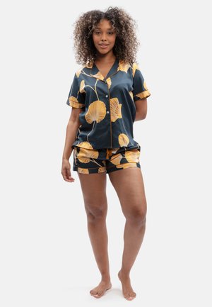 Woman standing barefoot wearing dark blue short-sleeve button shirt and shorts with large yellow floral print on a white background.