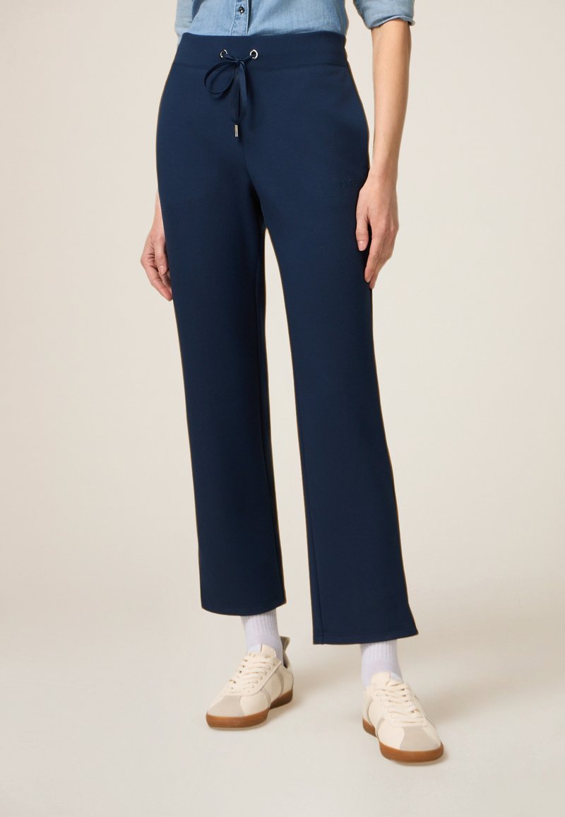 Pantalons de survêtement larges bleu marine en tissu doux, avec une taille élastique, un cordon de serrage et un petit logo sur le côté. Portés avec des baskets légères.