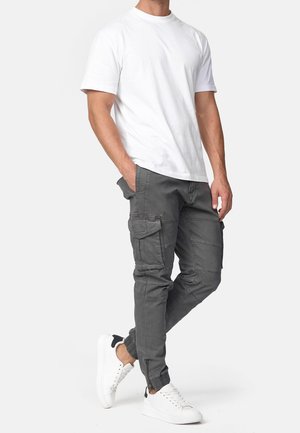 Mann trägt weißes T-Shirt mit kurzen Ärmeln, graue Cargohosen mit Taschen und weiße Sneakers mit schwarzen Akzenten, steht mit den Händen in den Taschen.