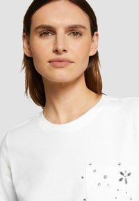 Witte t-shirt met een ronde halslijn en een decoratieve zak versierd met verschillende edelsteenaccenten. Het materiaal lijkt zacht en lichtgewicht te zijn.