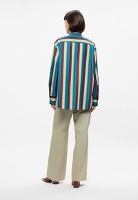 Ριγέ πουκάμισο σε μπλε, μωβ και κίτρινο χρώμα, με κουμπωτές μανσέτες και χαλαρή εφαρμογή, συνδυασμένο με ανοιχτό μπεζ παντελόνι σε φαρδιά γραμμή.