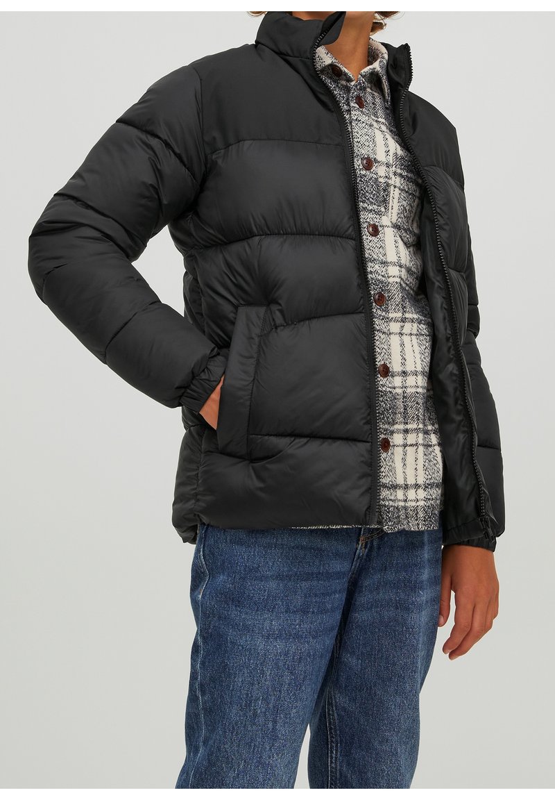 Osoba ubrana w czarną kurtkę puchową na białą i czarną koszulę w kratę z guzikami oraz niebieskie dżinsy, stojąca z jedną ręką w kieszeni.