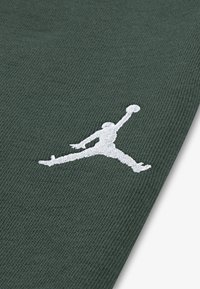 Zaļš audums ar izteiktu baltu izšūtu Jumpman logotipu. Gluda tekstūra bez redzamām raksturām vai akcentiem.