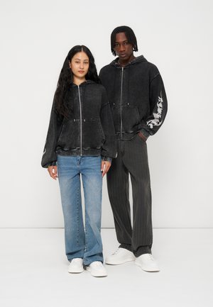 Deux personnes debout portant des sweat-shirts à capuche noirs avec fermeture éclair, l'un avec un jean bleu et l'autre avec un pantalon noir, toutes deux portant des baskets blanches.