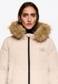 Chaqueta acolchada beige con capucha de piel sintética color marrón desmontable, cremallera negra y bolsillos laterales. Cuenta con un parche de logo en el pecho.