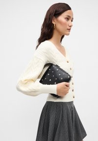 Pulover cremos cu tricotaj tip cablu și butoane aurii, asortat cu o fustă gri cu dungi subțiri. În mână se află o geantă mică neagră, cu ținte argintii.