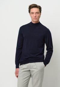Navyblauer Rollkragenpullover, gestrickte Textur, lange Ärmel, gerippte Bündchen und Saum, kombiniert mit hellgrauen maßgeschneiderten Hosen.