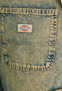 Overoles de trabajo de mezclilla con un bolsillo trasero prominente que tiene un parche rectangular con el logotipo "Dickies" en rojo y azul.