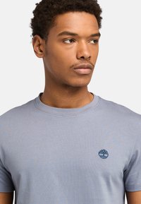 Ung mand med kort krøllet hår, der ser til siden, iført en lyseblå t-shirt med et lille broderet trælogo på brystet.
