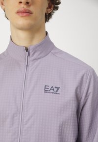 Svijetloplavi zip-up sakou s teksturiranim uzorkom. Ima stojeći ovratnik i logo EA7 Emporio Armani na prsima.