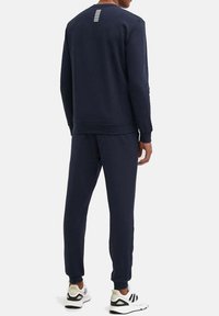 Felpa blu navy e pantaloni jogger coordinati in tessuto. La felpa presenta un dettaglio a righe sul retro; i pantaloni hanno polsini e vita elastici.