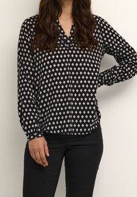 Frau in schwarzer Bluse mit langen Ärmeln und weißem Diamantmuster sowie schwarzen Hosen, die vor einem schlichten hellen Hintergrund steht.