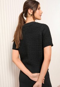 Melnā īsā piedurkne džemperis ar strukturētu, audumu rakstu; ribotas aproces un apakšmalas; brīva piegriezuma. Modeļa mati sasieti zirgaste, redzami aiz muguras.