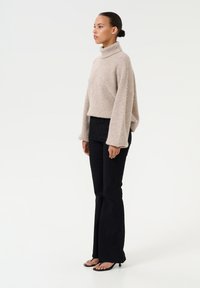 Maglione beige con collo a imbuto e texture a coste, abbinato a jeans neri a palazzo e sandali con tacco neri. Modella in posa di profilo.