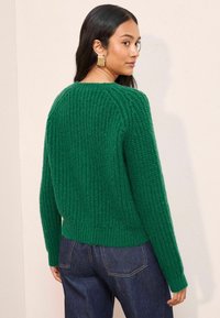 Zelený pletený svetr s žebrovanou texturou, kulatým výstřihem a přiléhavým lemem. Ve spojení s tmavě modrými džíny, které vytvářejí uvolněnou siluetu.