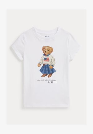 Bela bomba majica iz bombaža, ki vsebuje ilustriranega medveda v puloverju z ameriško zastavo, modra jeans krila in rjave škornje. Podpis spodaj.