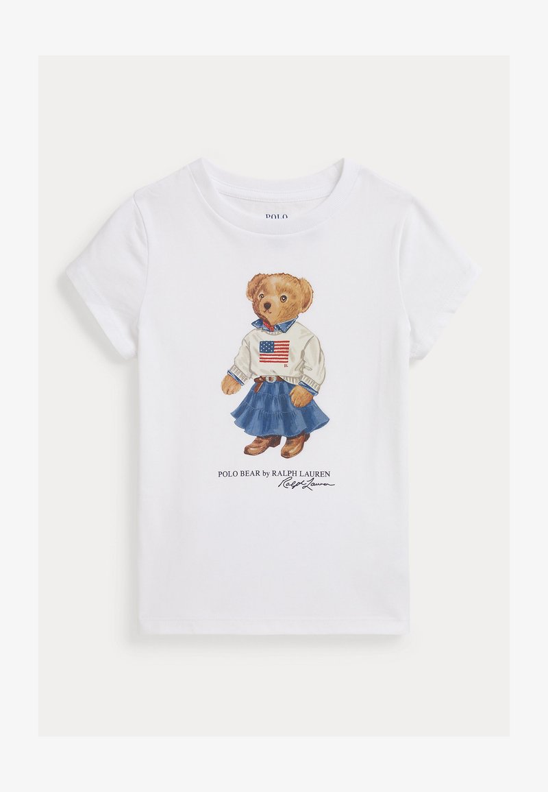 Balts kokvilnas T-krekls ar ilustrētu lāci džemperī ar Amerikas karogu, zila denima svārki un brūni zābaki. Paraksts zem attēla.