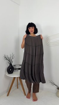 Femme avec une coupe de cheveux au carré noir tenant une jupe sombre à volants, debout pieds nus à côté d'une table ronde blanche avec des livres et un vase noir.