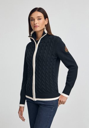 Gebreide marineblauwe trui met een kabelpatroon, hoge kraag, crèmekleurige rits en geribbelde manchetten. Heeft een logopatch op de linkerarm.