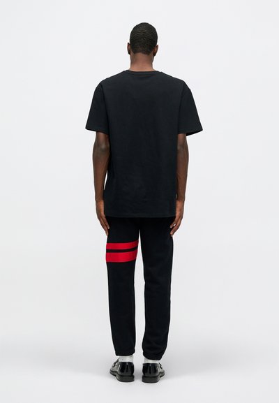 Juoda medvilninė marškinėliai su laisvu siluetu, derinami su juodais sportiniais džinsais, ant kairiojo blauzdos turinčiais raudonas horizontalias juostas. Išvaizdą užbaigia juodi batai.