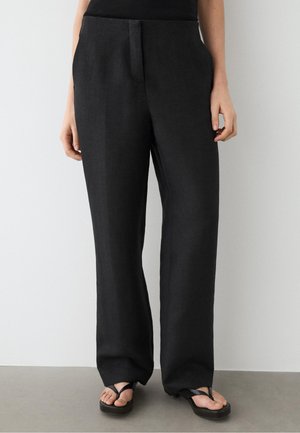 Femme portant un pantalon noir ample avec des poches latérales et des sandales plateformes noires sur un fond clair uni.