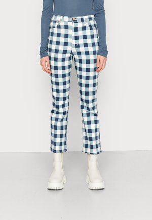 Person iført blå og hvide ternede bukser, en blå bluse med lange ærmer og hvide chunky ankelstøvler, stående mod en ensfarvet baggrund.