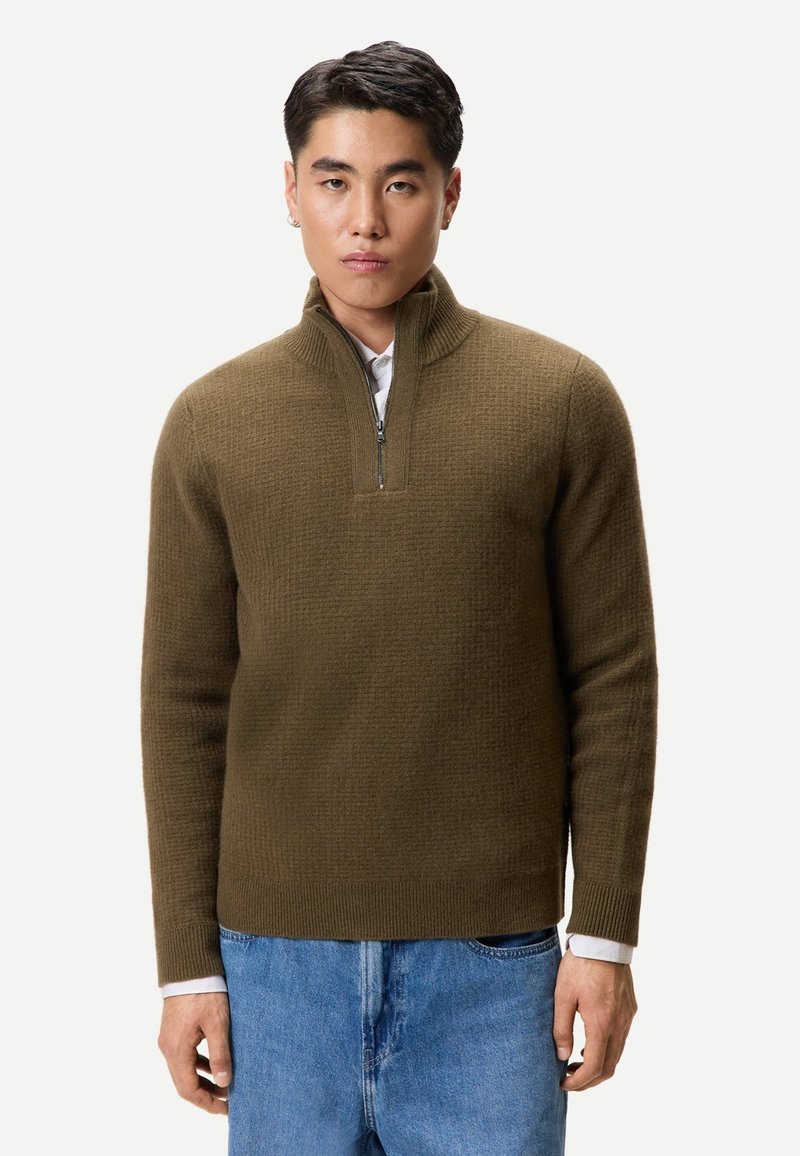 Junger Mann trägt einen olivgrünen Pullover mit Reißverschluss bis zur Brust über einem weißen Hemd und blauen Jeans, steht vor einem schlichten weißen Hintergrund.