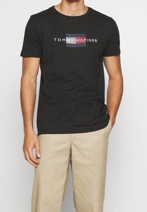 Mężczyzna w czarnej koszulce Tommy Hilfiger z logo i beżowych spodniach, stojący z jedną ręką w kieszeni na gładkim tle.