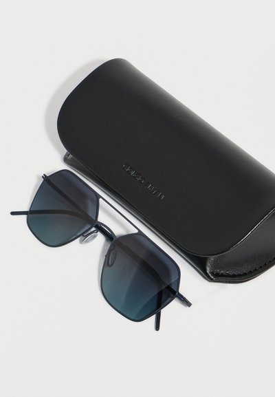 Μαύρα γυαλιά ηλίου με τετράγωνο πλαίσιο και σκούρους φακούς με διαβάθμιση, τοποθετημένα δίπλα σε μια μαύρη δερμάτινη θήκη Giorgio Armani πάνω σε λευκή επιφάνεια.