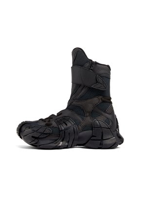 Bota atlética negra con un upper de malla texturizada, suela de goma y correa ajustable. Presenta un diseño de zigzag prominente en el costado.