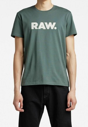 Osoba nosząca zielony T-shirt z napisem „RAW.” dużymi białymi literami, w połączeniu z czarnymi spodniami, stojąca na jednolitym tle.