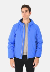 Uomo che indossa una giacca con cappuccio blu brillante, aperta sopra un maglione grigio e pantaloni neri, in piedi di fronte a uno sfondo bianco semplice.