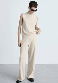 Beige gestricktes, ärmelloses Oberteil mit hohem Kragen, kombiniert mit passenden weit geschnittenen Hosen. Das Model trägt schwarze rechteckige Sonnenbrillen und Schuhe.