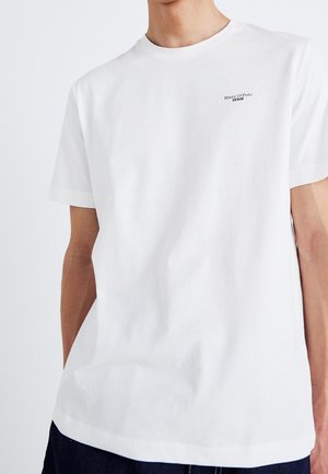 Młody dorosły noszący gładką białą koszulkę z krótkim rękawem z małym logo "Marc O’Polo Denim" na lewej piersi, w zestawieniu z ciemnymi spodniami.