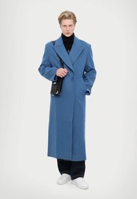 Dlhý modrý kabát zo zmesi vlny, s výstrihom do V, jednobodovým zapínaním a širokými rukávmi. V kombinácii s čiernymi nohavicami a kabelkou.