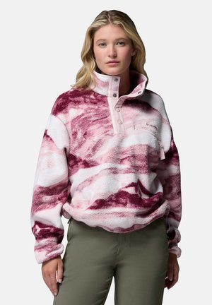 Donna con capelli biondi indossa un pullover in pile rosa e bianco con motivo a fantasia, colletto con bottoni automatici e pantaloni verde oliva, in piedi davanti a uno sfondo uniforme.