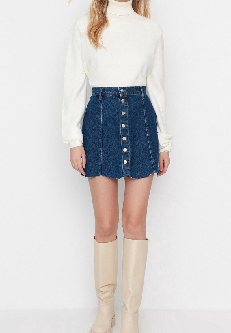 Denim mini-rok met een licht uitlopende zoom, sluiting met knopen aan de voorkant en vijf zakken. Draagt een witte lange mouwen coltrui en beige laarzen.