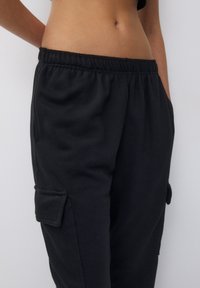 Pantalon cargo noir avec une taille élastique, des poches latérales et une texture douce en coton. Il présente une coupe ample et des détails de poche cousus.