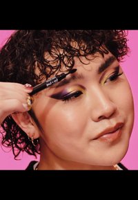 Crayon à sourcils noir appliqué sur le sourcil d'un mannequin. Présente un maquillage des yeux violet et jaune éclatant, des lèvres brillantes et des cheveux bouclés texturés.