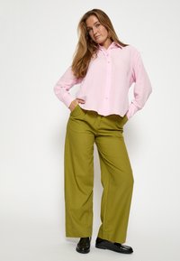 Roza majica z dolgimi rokavi in gumbi spredaj, v kombinaciji z olivno zelenimi širokimi hlačami. Črni čevlji preproste zasnove in gladke teksture.