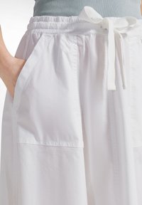 Hvide, løstsiddende bukser med lommer og et stofbælte, en persons hånd afslappet placeret i en lomme.